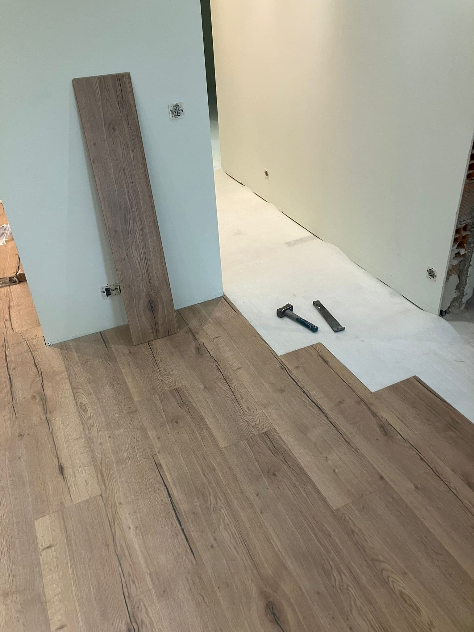 Piso laminado de madeira sendo instalado sobre manta branca com martelo e barra de tração.